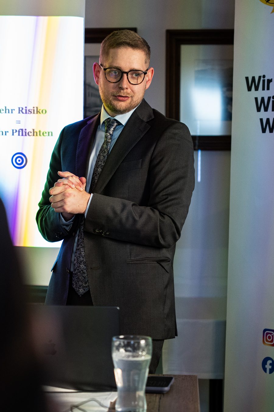 Vortrag vor einer Leinwand mit der Folie „Mehr Risiko = mehr Pflichten“, daneben ein Roll-up-Banner